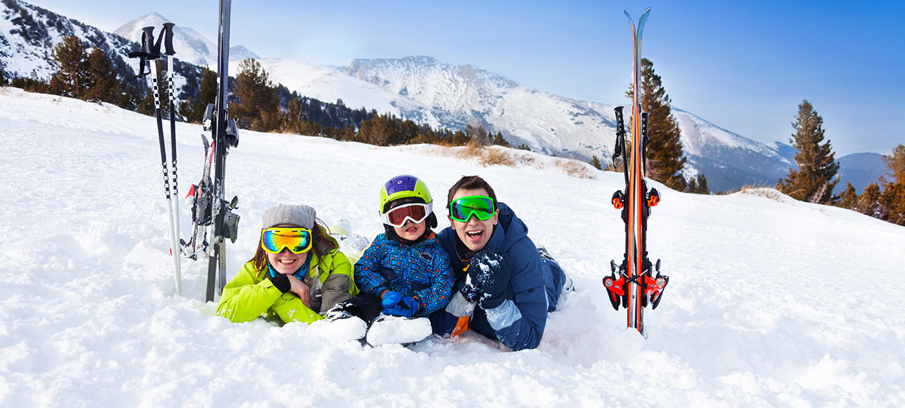 Famiglia con attrezzatura da sci, caschi e occhiali sdraiata in mezzo alla neve e sorridente